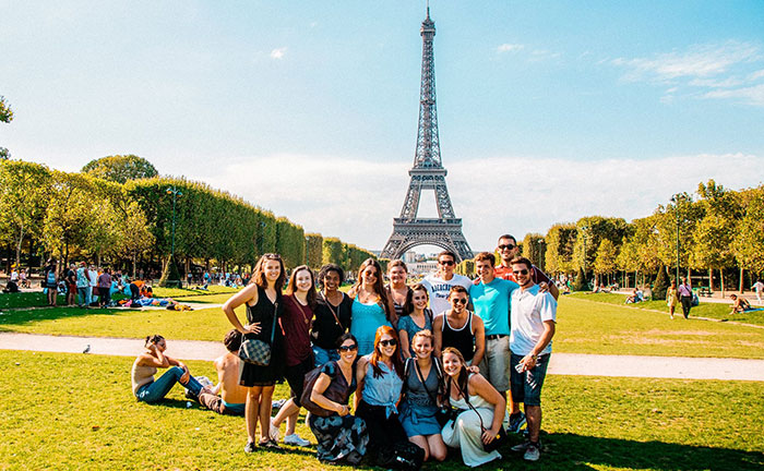 Students smiling for a group photo on a beautiful day at the Eiffel Tower with bright green grass and blue skies